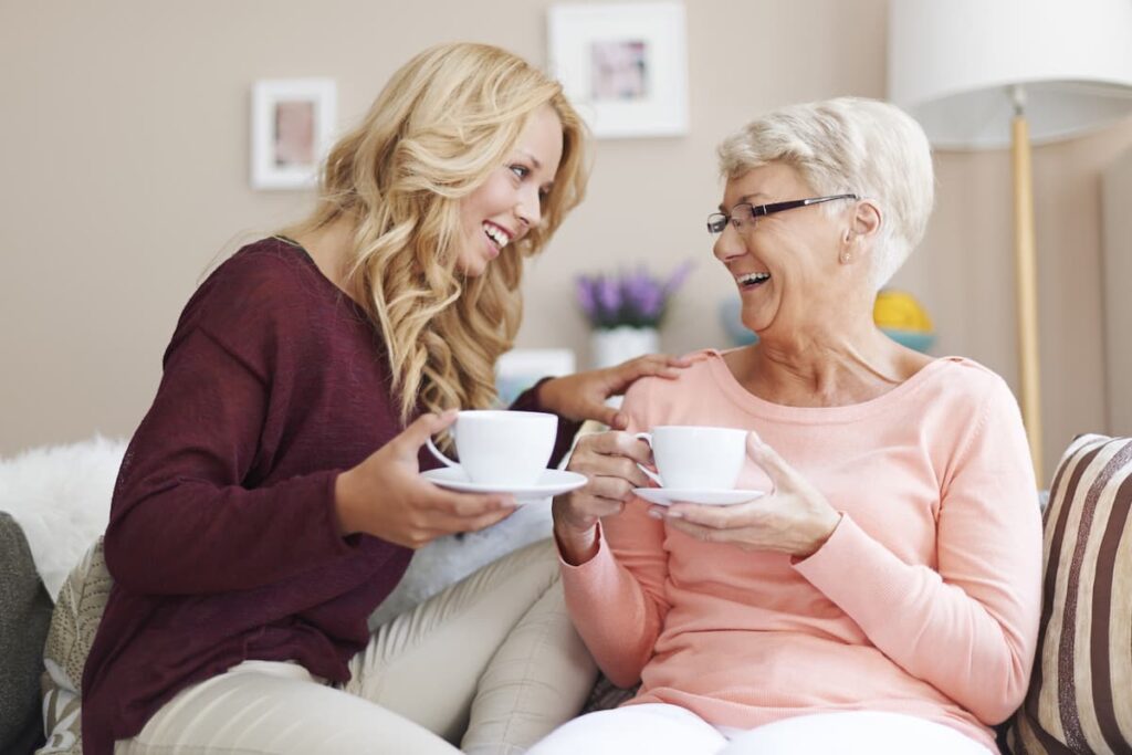 Prayers For Grandmas - grandmother and daughter sitting on the couch drinking tea together at smiling at each other