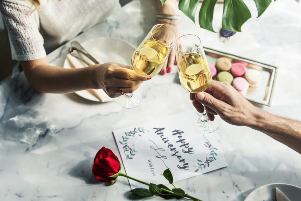 Anniversary Prayers - couple clinking champagne glasses together at the dinner table that has macarons, a red rose, and a sign saying "Happy Anniversary"
