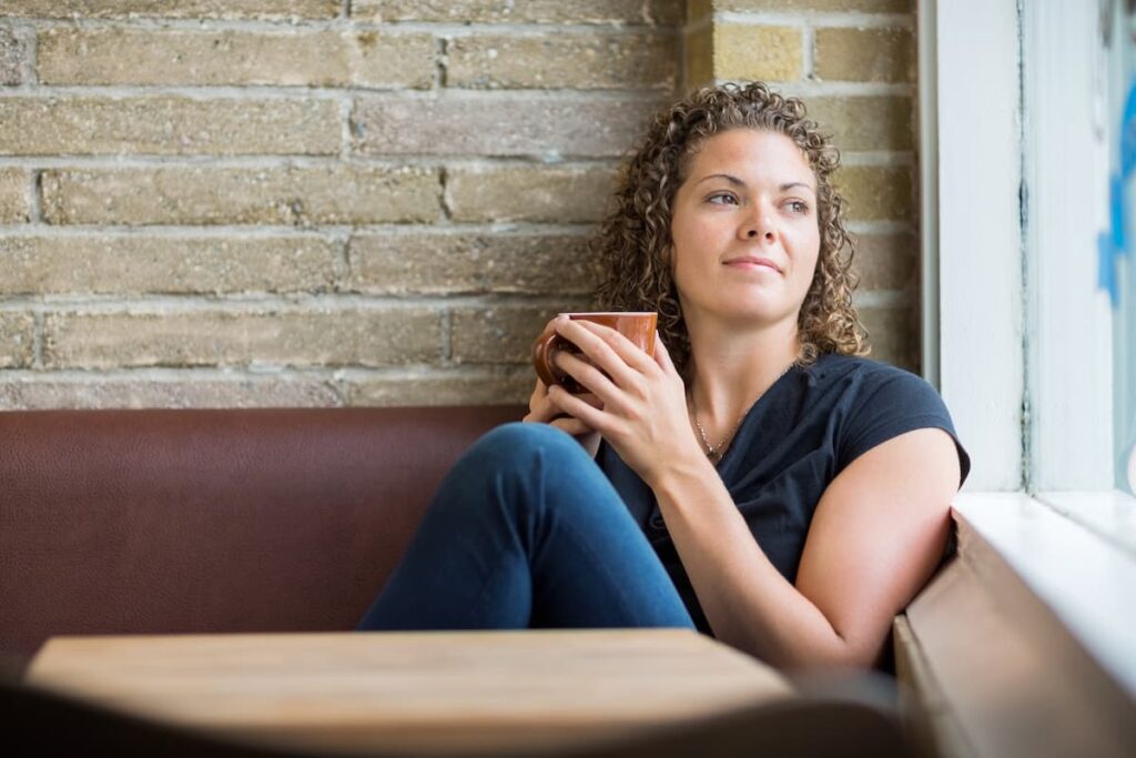 Prayers For Discernment - woman sitting in booth holding a mug of coffee looking out the window with a slight smile