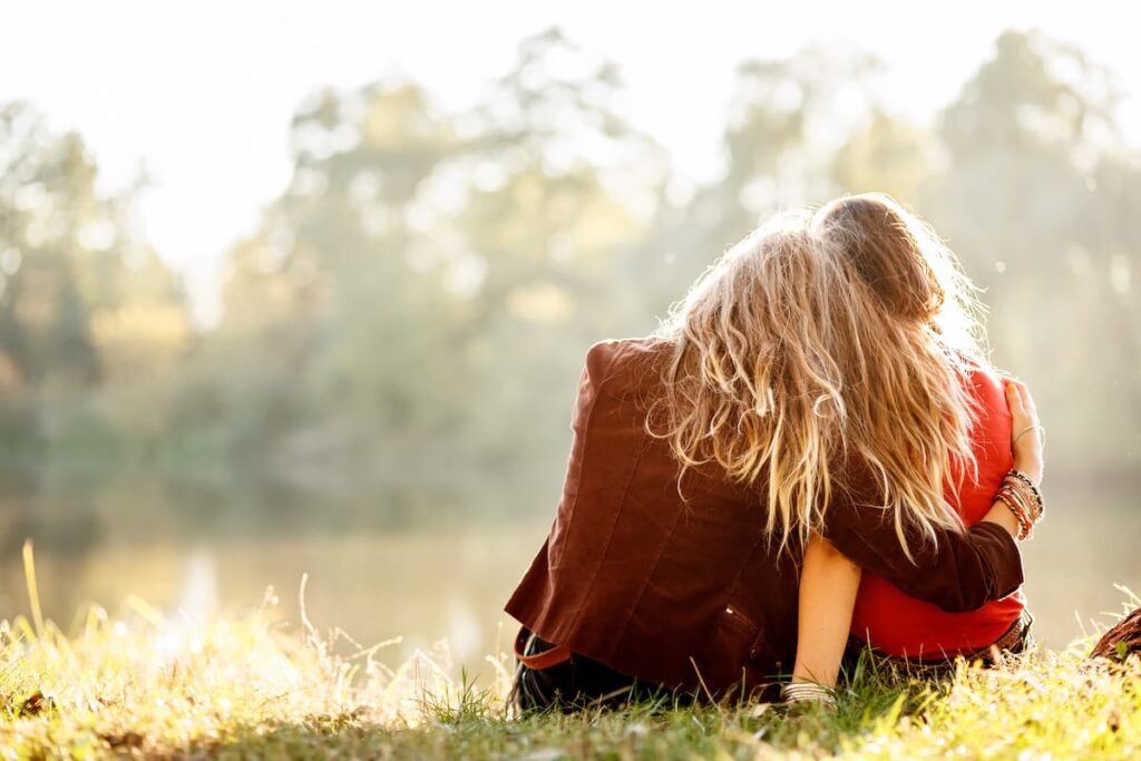 Prayers For Unsaved Loved Ones - woman sitting in the grass, hugging the woman sitting next to her, facing away from the camera