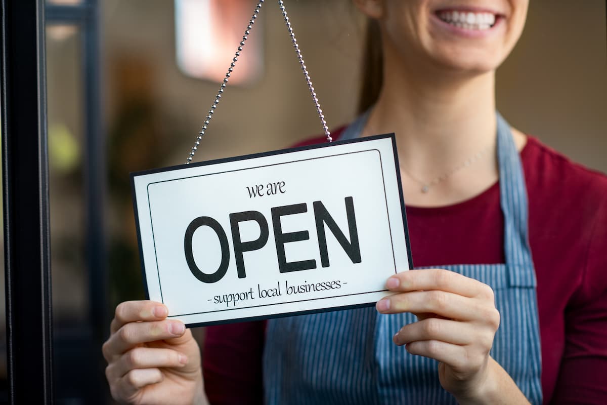 4 Business Prayers For Success - Woman smiling turning front door  Closed sign to Open 