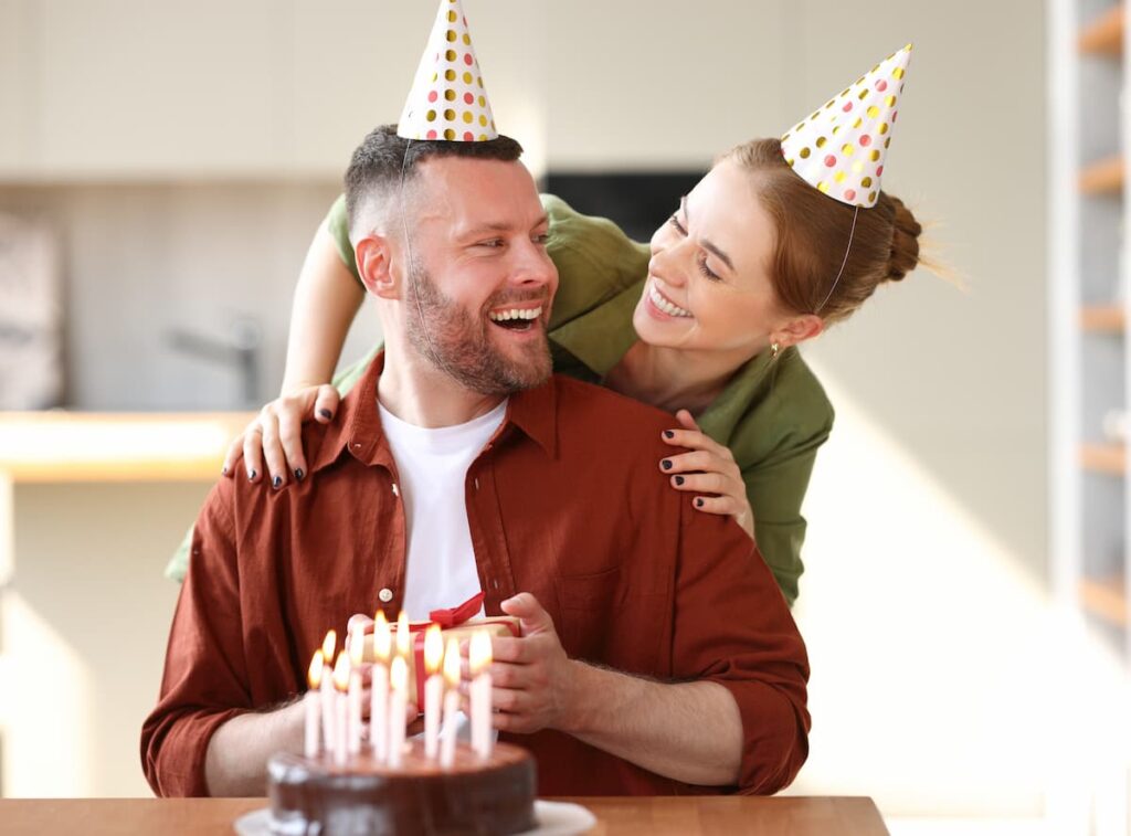 Wife wearing a party hat surprising husband with a birthday cake and gift