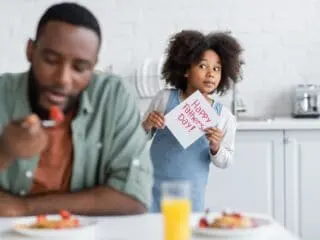 Bible verses for Father's Day - little girl sneaking a Father's Day card to her dad who is eating breakfast