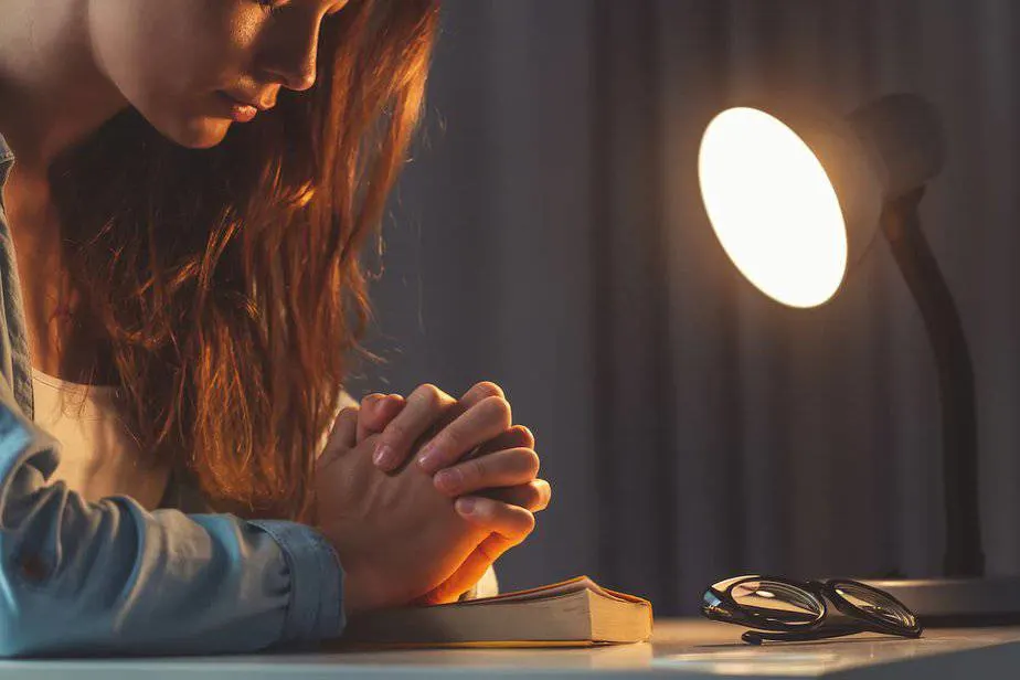 young woman praying with hands clasped on a bible at night for 12 Bible Verses For Nighttime Prayer