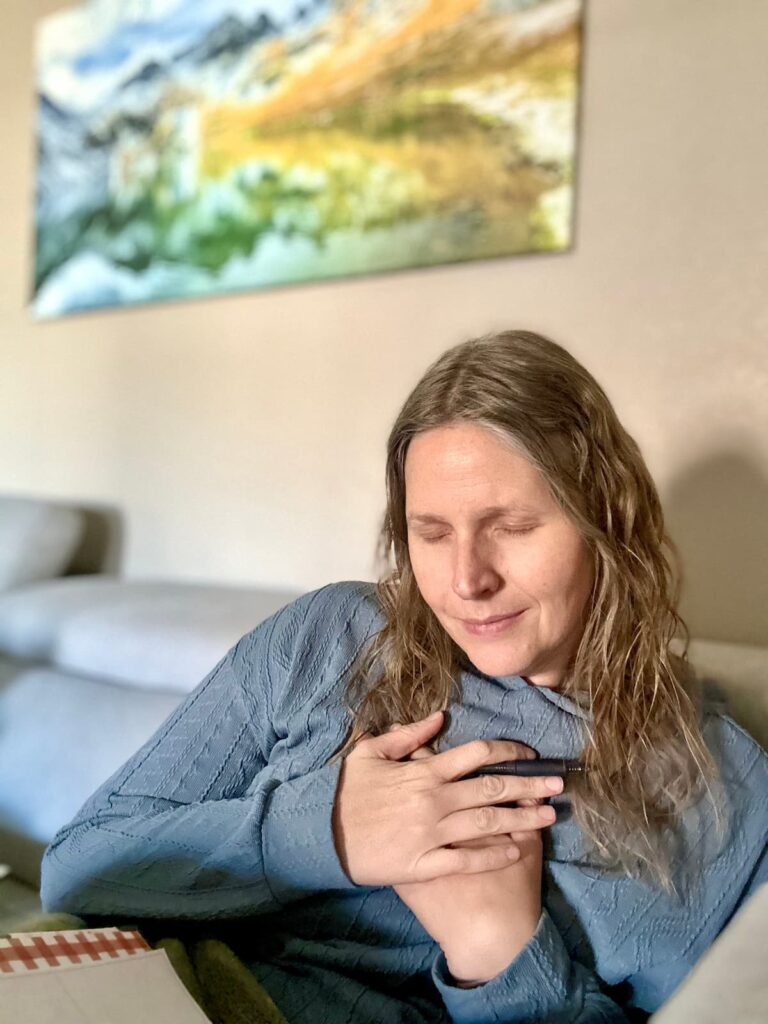 Woman sitting on her couch with her eyes closed hands gently folded over her heart, and eyes closed with a slight smile on her face as she prays