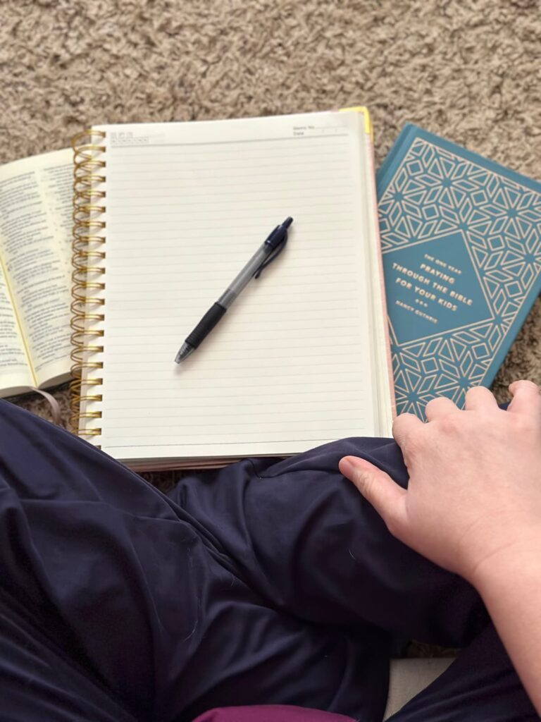 Overhead picture of crossed legs and a hand on their leg with a blank pag in an open journal topped with a pen and an open Bible and prayer book for children on the floor next to the notebook