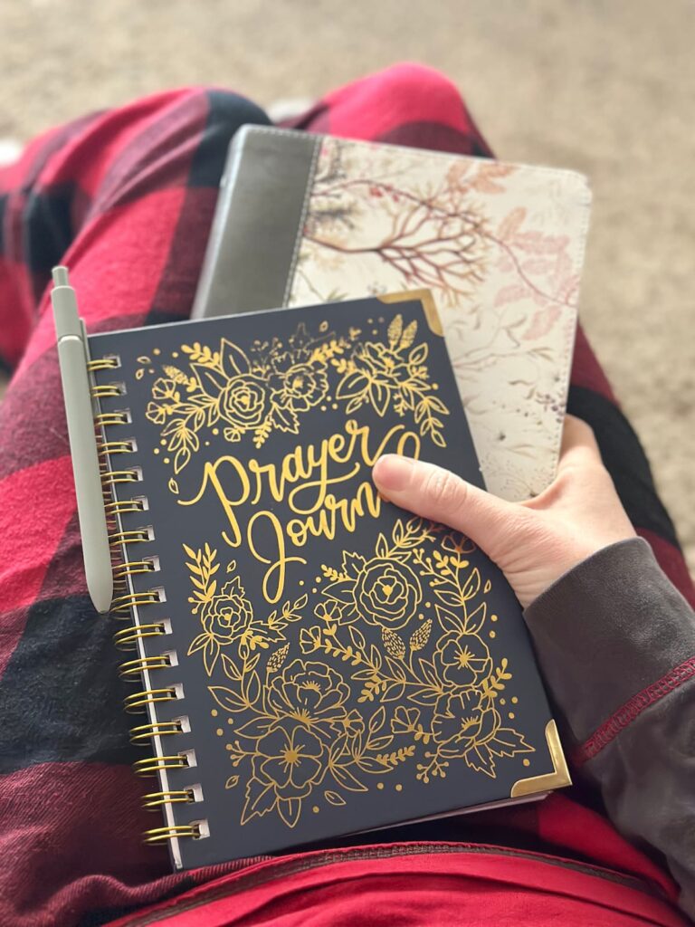 Woman sitting on the floor with bent legs wearing red flannel pj pants holding a Bible and a prayer journal spiral notebook and pen in her lap