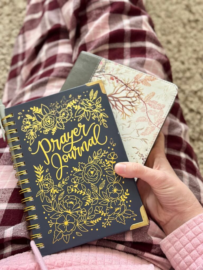 Prayer journal spiral notebook with a Bible held by a woman sitting on the floor with her legs outstretched with a pink sweatshirt and pink flannel pj pants