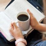 Open Bible in lap with hands holding a mug of coffee just above it